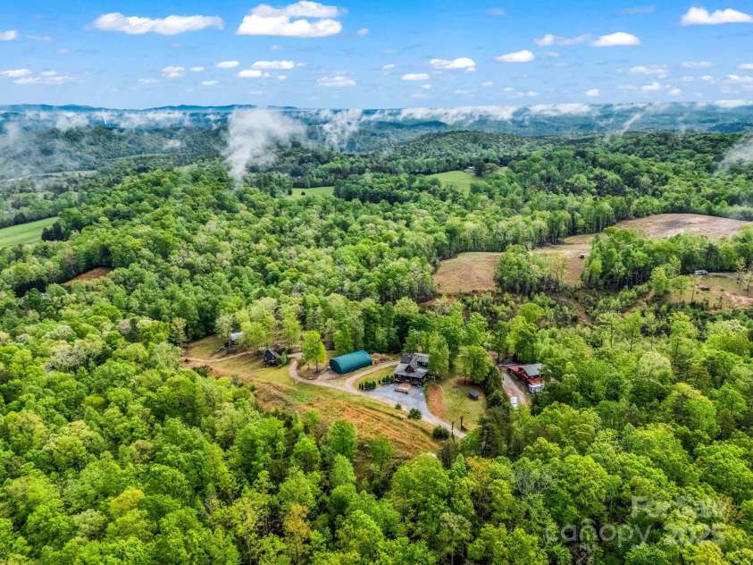 1002 CC Road, Rutherfordton, NC 28139.  MLS# CAR4249234, YatesRealty ID 13761. Aerial view of main cabin (center with circular drive), guest cabin (far left behind the trees), metal arch storage building (green), and barn/workshop (red) far right.