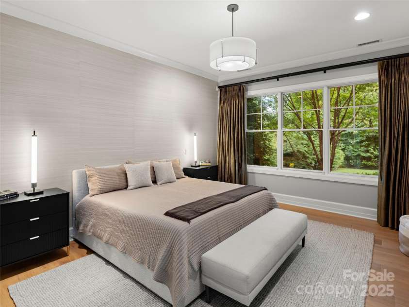 62 Running Creek Trail, Arden, NC 28704.  MLS# CAR4299725, YatesRealty ID 1785. Tranquil primary suite that looks out to private and beautiful backyard landscaping. Newer grass cloth wall covering accent on wall behind bed. Wide plank hardwood floors. Lush silk draperies convey with the house. 