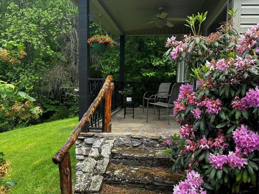 51 Atari Road, Waynesville, NC 28786.  MLS# CAR4256064, YatesRealty ID 23630. Front porch surrounded by flowers and flowering bushes with a hand carved railing.