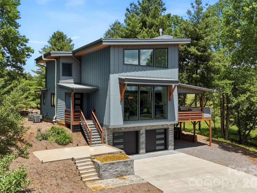 22 Lara Drive, Mills River, NC 28759.  MLS# CAR4382358, YatesRealty ID 23687. Angled exterior view showcasing the home’s sleek modern architecture, elevated covered deck, and thoughtfully designed entry staircase. Surrounded by lush greenery for added privacy and natural beauty.