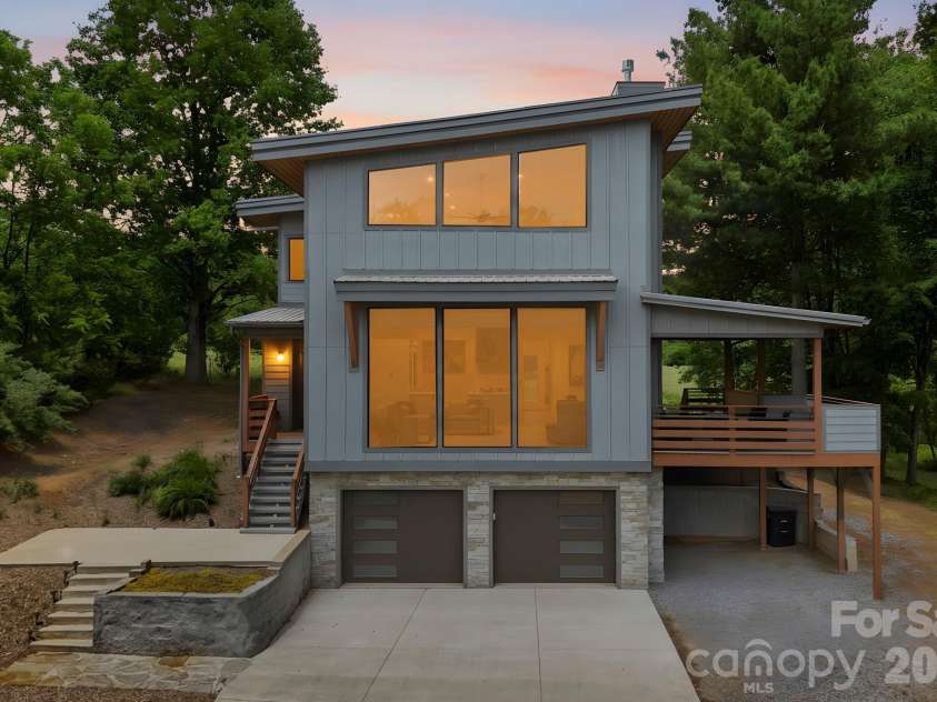 22 Lara Drive, Mills River, NC 28759.  MLS# CAR4382358, YatesRealty ID 23687. Striking modern mountain home with dramatic floor-to-ceiling windows, clean architectural lines, and warm interior lighting that creates a stunning evening presence. The covered side deck and wooded setting add to the home