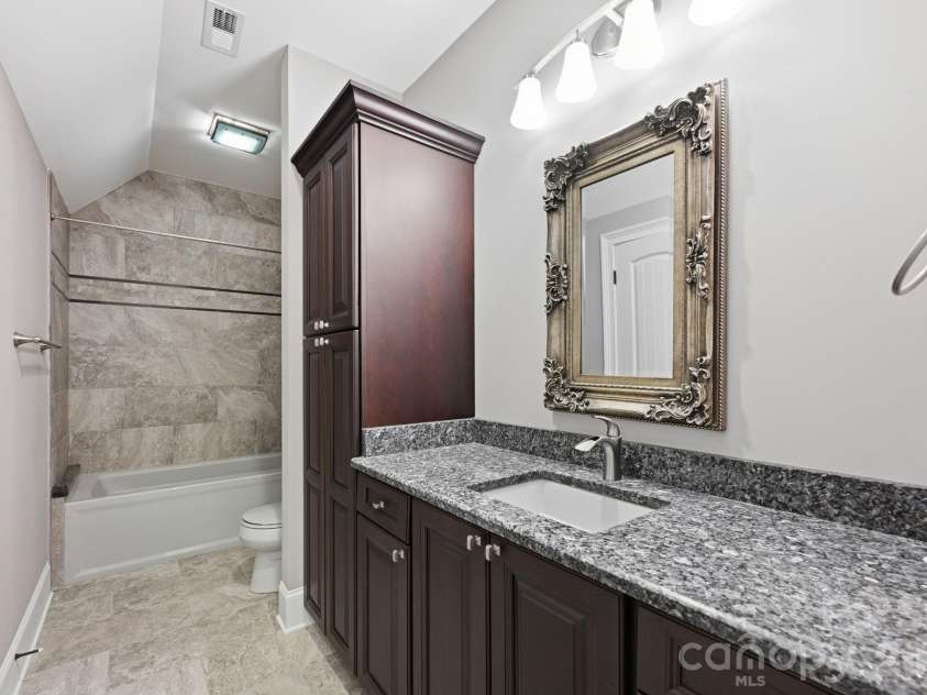 108 Washam Road, Mooresville, NC 28117.  MLS# CAR4334255, YatesRealty ID 4048. Modern bathroom featuring granite countertops, custom dark wood cabinetry, and a framed decorative mirror with overhead lighting. The space includes a tiled tub/shower combination and neutral finishes, creating a clean an