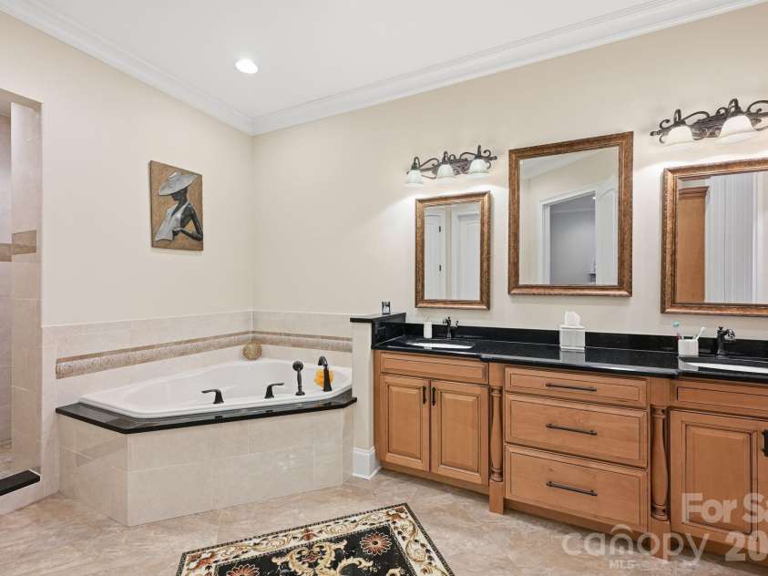 108 Washam Road, Mooresville, NC 28117.  MLS# CAR4334255, YatesRealty ID 4048. Spacious primary bathroom featuring a soaking tub with tile surround, double vanity with granite countertops, and framed mirrors with decorative lighting. The room offers a clean, elegant design with tile flooring and a b
