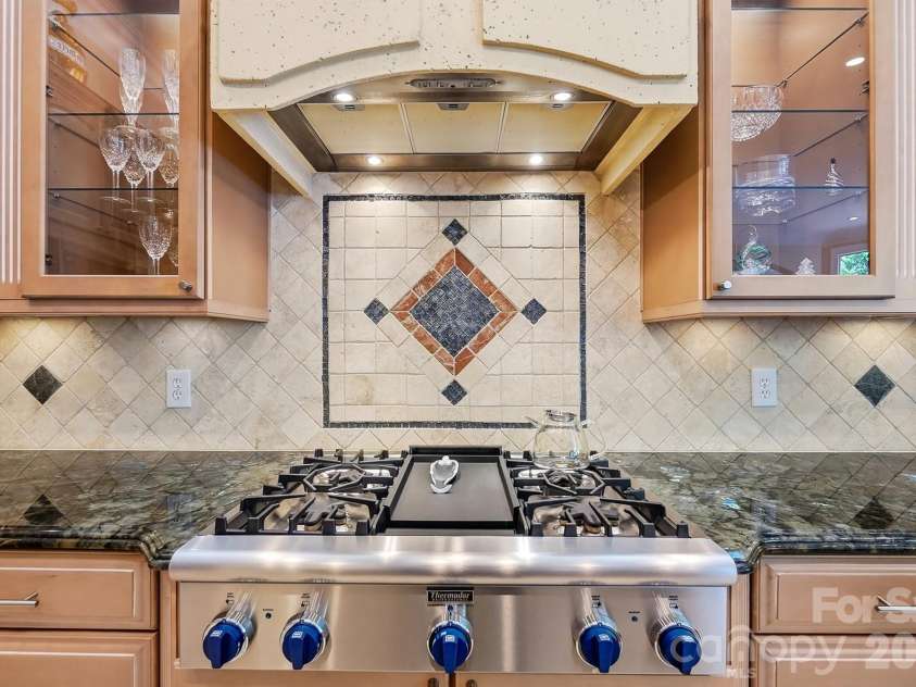 20124 Bascom Ridge Drive, Cornelius, NC 28031.  MLS# CAR4408389, YatesRealty ID 44623. The kitchen’s professional-style range creates a striking focal point, framed by illuminated glass-front cabinetry, a substantial custom hood, and decorative tilework. Rich stone counters and thoughtful detailing 