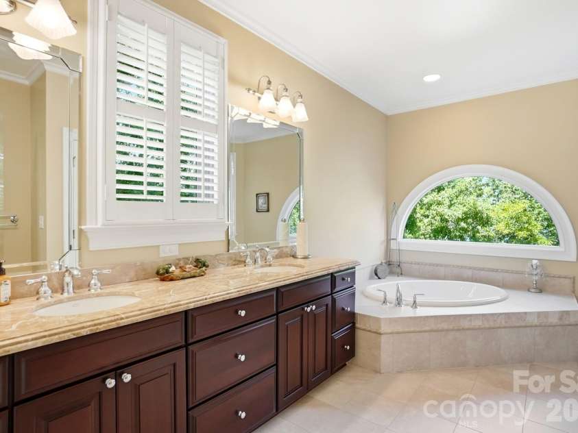 20124 Bascom Ridge Drive, Cornelius, NC 28031.  MLS# CAR4408389, YatesRealty ID 44623. The private bath features a long dual-sink vanity with rich cabinetry and stone countertops, along with a spacious soaking tub positioned beneath a distinctive arched window. Natural light and leafy views give the