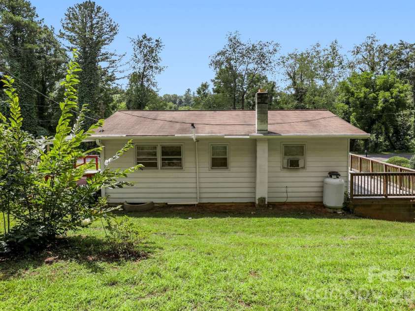 27 McGee Circle, Fairview, NC 28730.  MLS# CAR4415904, YatesRealty ID 45673. Rear exterior view showcasing the home’s grassy yard, trees, and deck access.